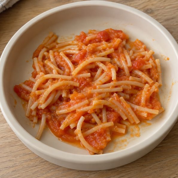 Rövidre vágott spagetti paradicsomos-répás szósszal, világos babatányérban, faasztalon.