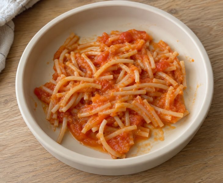 Rövidre vágott spagetti paradicsomos-répás szósszal, világos babatányérban, faasztalon.